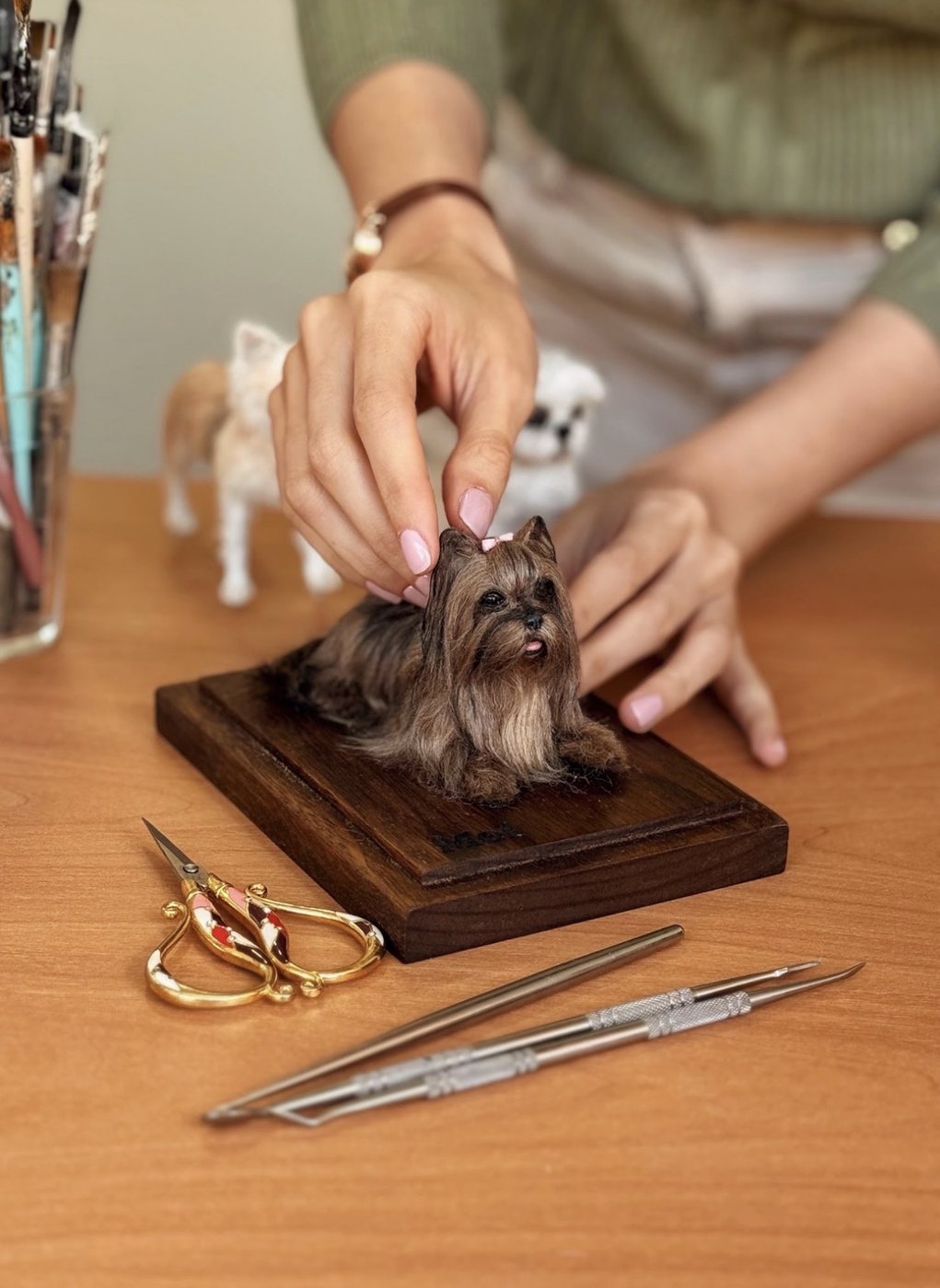 Jiceli finalizando uma miniatura realista de cachorro no ateliê, com tesoura e ferramentas de modelagem